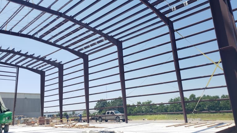 Tall red-iron columns and rafters with horizontal wall girts on a new concrete slab, crew and pickup truck in the background