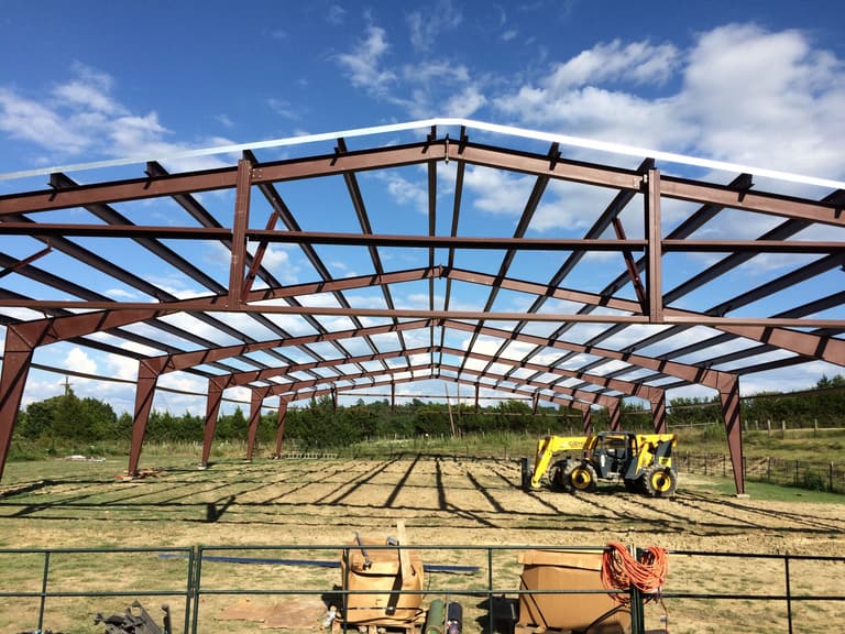 Red-iron gable frames and purlins of a large steel building on a rural lot, with a yellow telehandler parked inside the frame